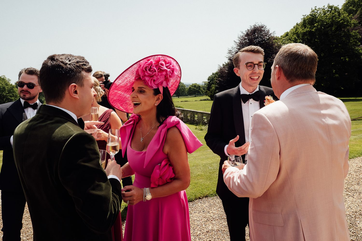 Elegant wedding guests in formal attire, including a woman in a pink dress and fascinator, socializing outdoors.