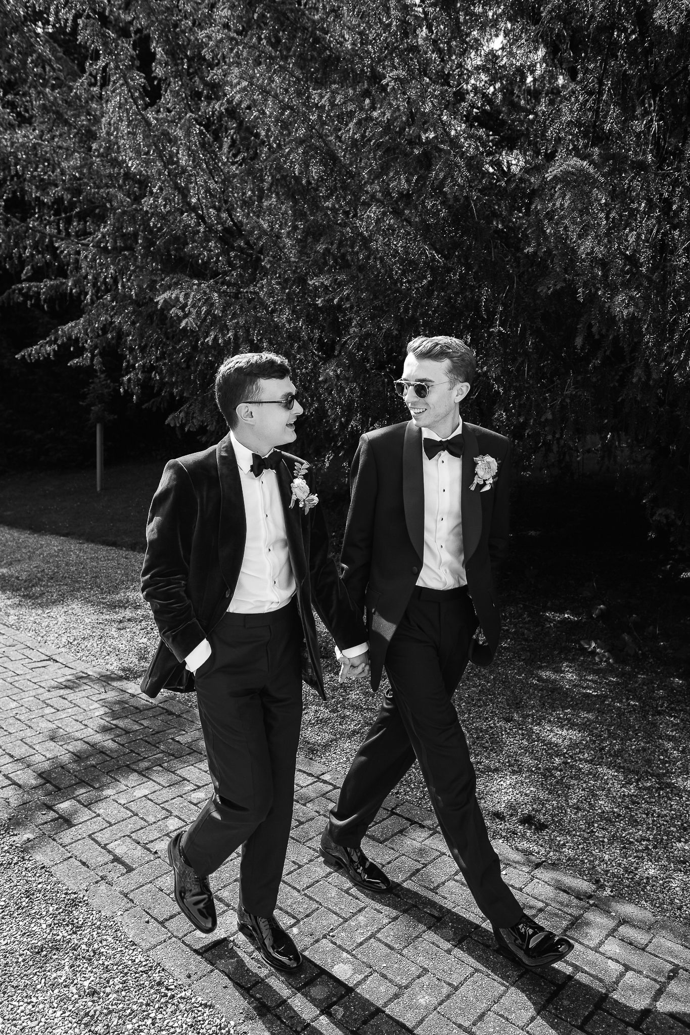 Black and white photo of two grooms in tuxedos holding hands and walking outdoors on their wedding day.