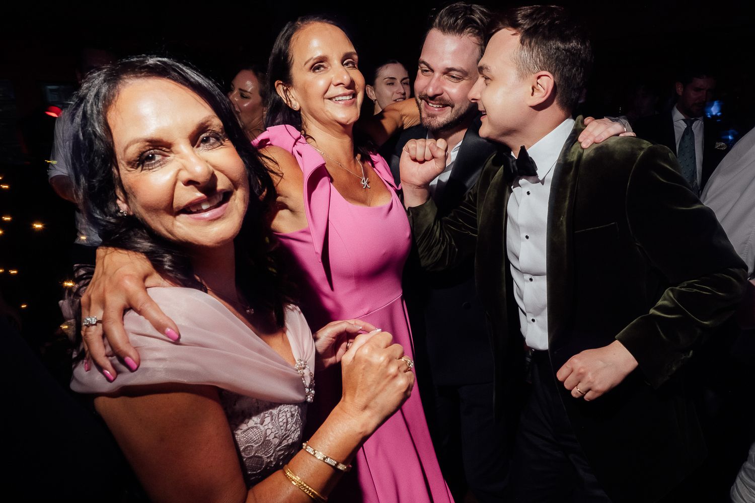 Guests dancing and laughing joyfully at a wedding reception, wearing elegant formal attire including a pink dress and velvet suit.