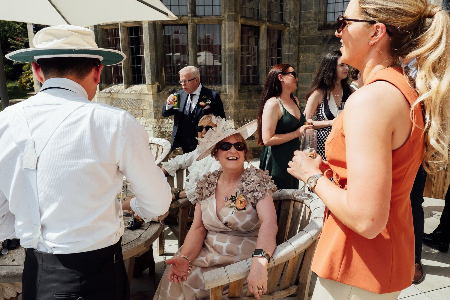 Guests in elegant attire chat outdoors at a sunny wedding reception, with a woman in a floral hat seated and smiling.