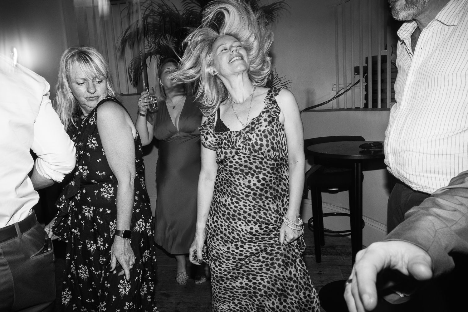 Black and white photo of women dancing at a lively party, one with windswept hair in a leopard print dress, surrounded by guests.