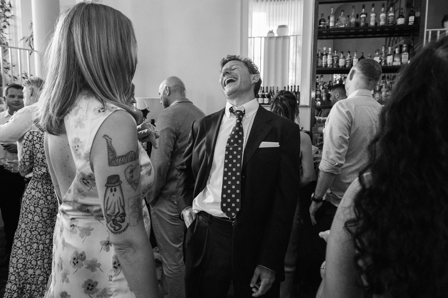 Black and white photo of a laughing man in a suit and polka dot tie at a lively indoor party, beside a woman with arm tattoos.
