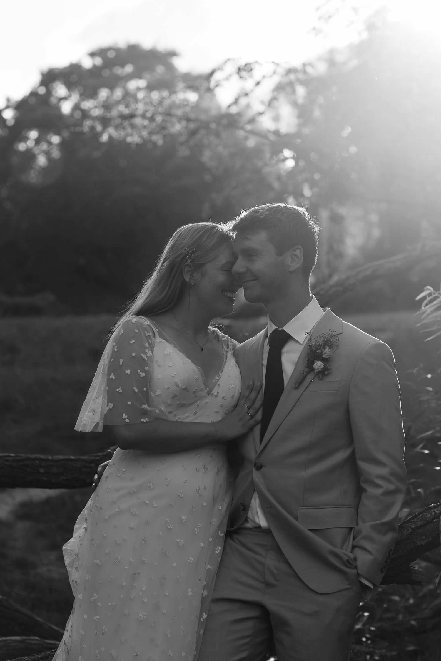Black and white photo of a bride and groom touching foreheads and smiling in a sunlit garden during their outdoor wedding.