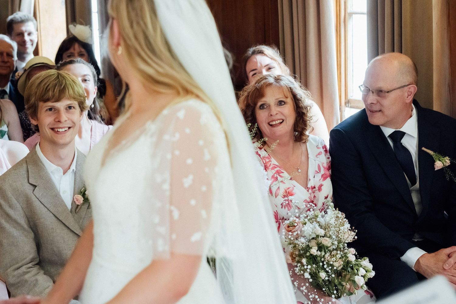Bride in a white dress and veil faces smiling wedding guests, including a woman holding a bouquet and a man in glasses, during the ceremony.