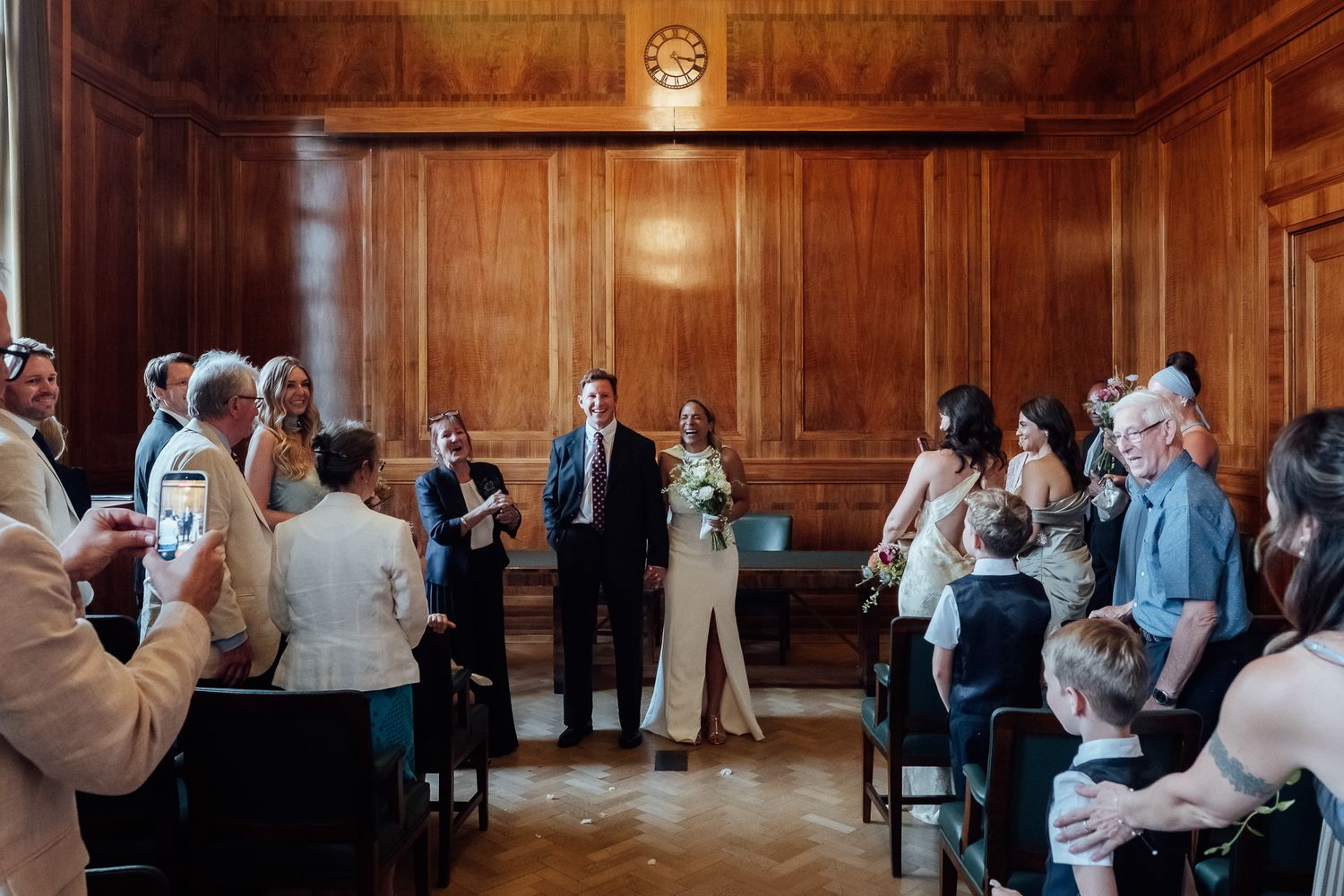 Bride in a white dress holding a bouquet stands beside a groom in a dark suit as guests watch in a wood-paneled wedding ceremony room.