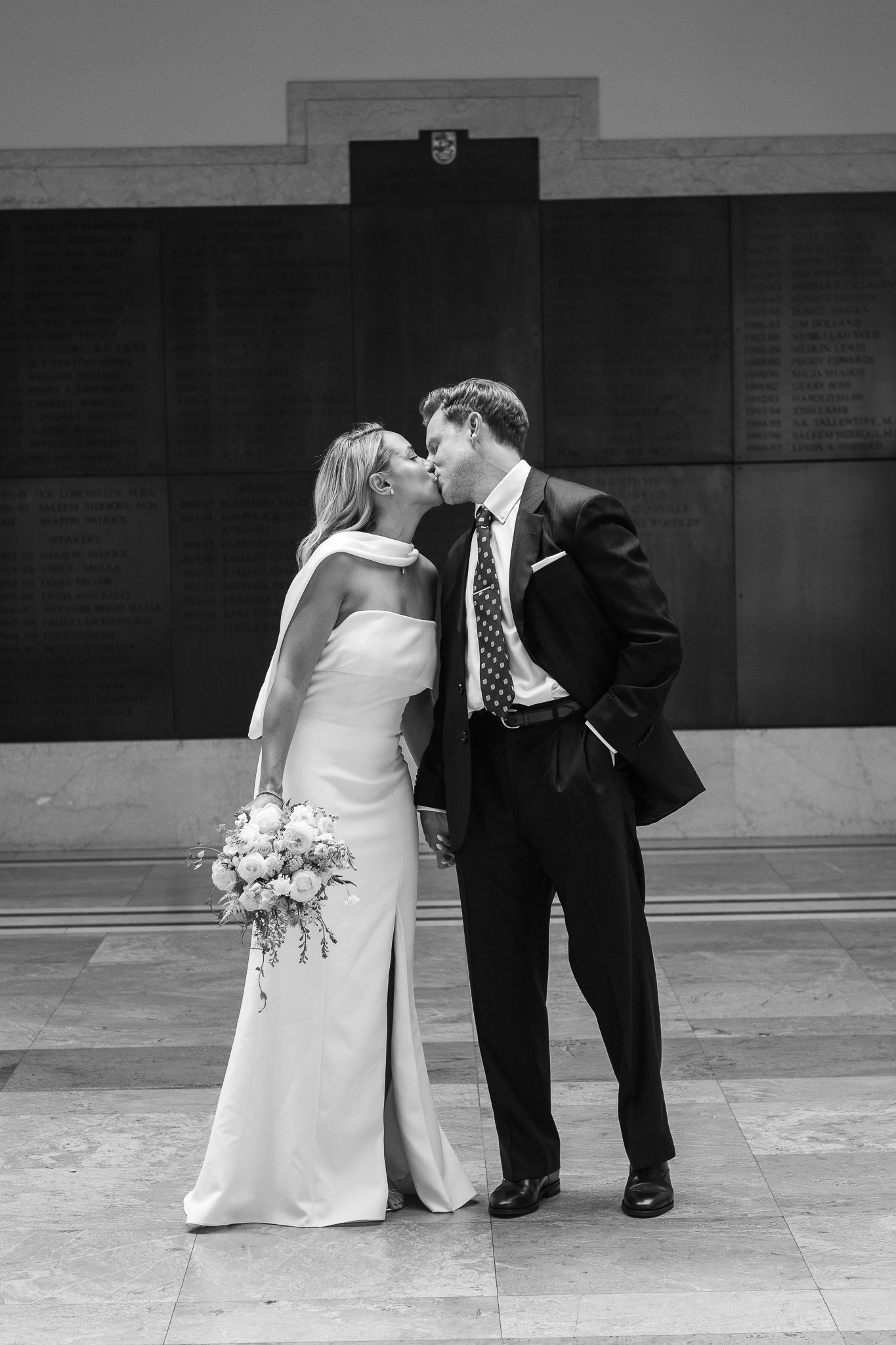 Black and white photo of a bride in a sleek strapless gown holding a bouquet, kissing her groom in a dark suit inside a stately marble hall.