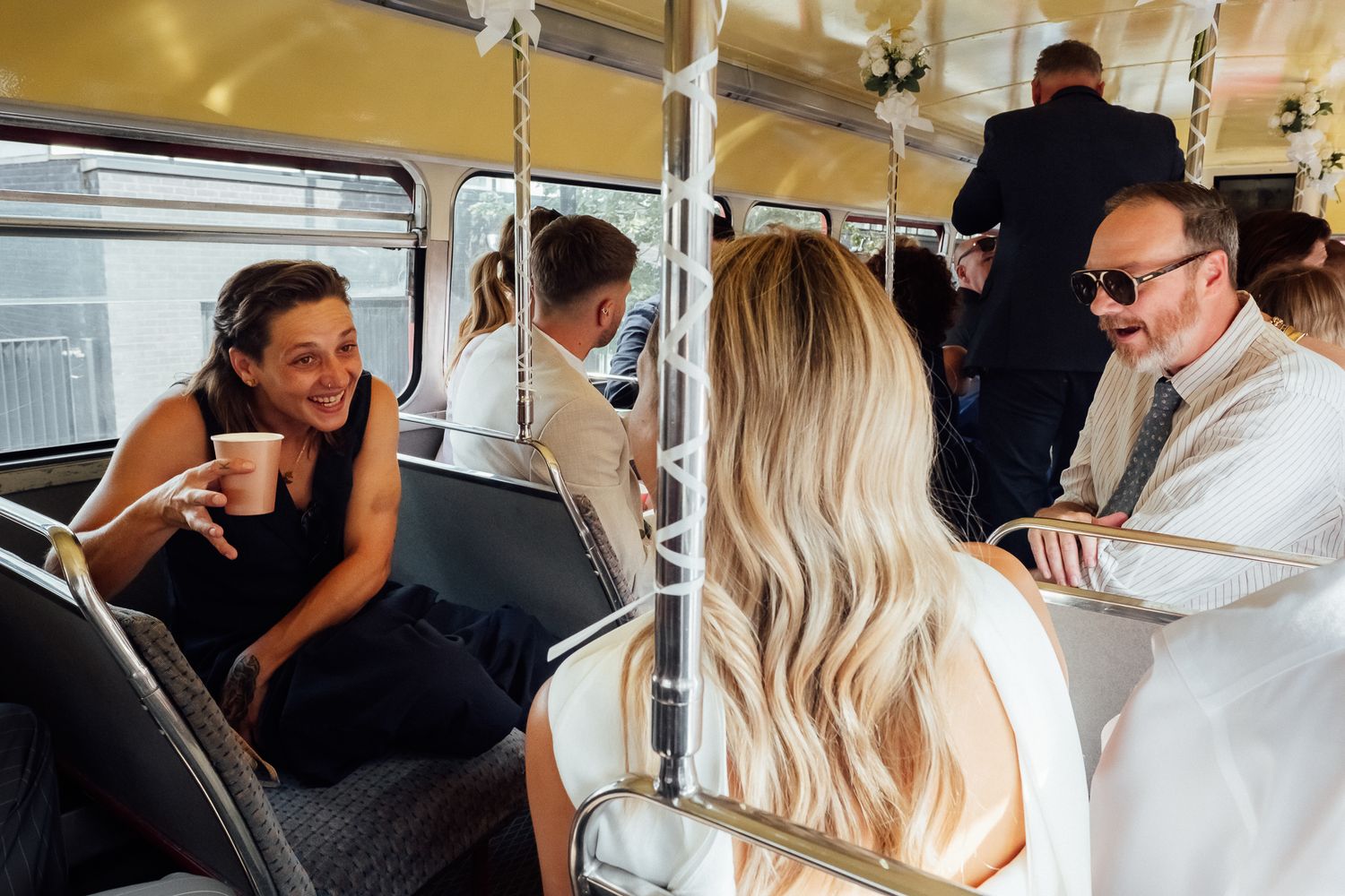 Smiling woman holding a cup chats with fellow passengers on a vintage-style bus decorated with flowers during a festive group ride.
