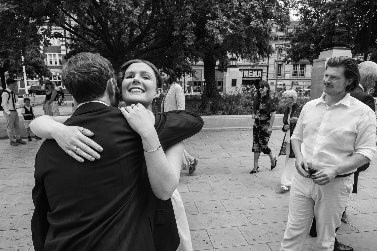 A smiling woman embraces a man in a dark suit in a sunny tree-lined plaza while guests mingle nearby at an outdoor wedding celebration.