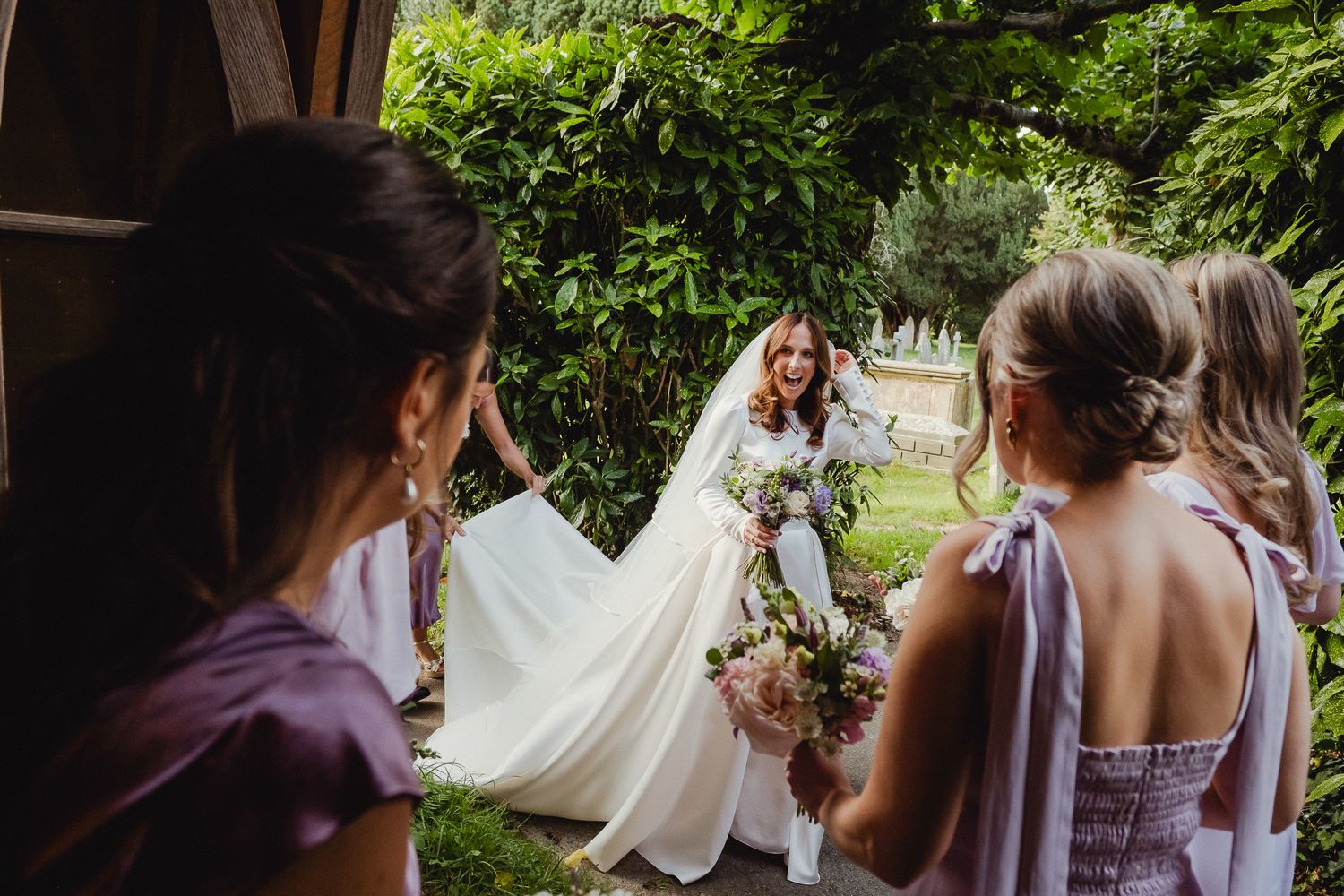 A wedding party walks through a lush garden setting while holding bouquets and wearing purple dresses.