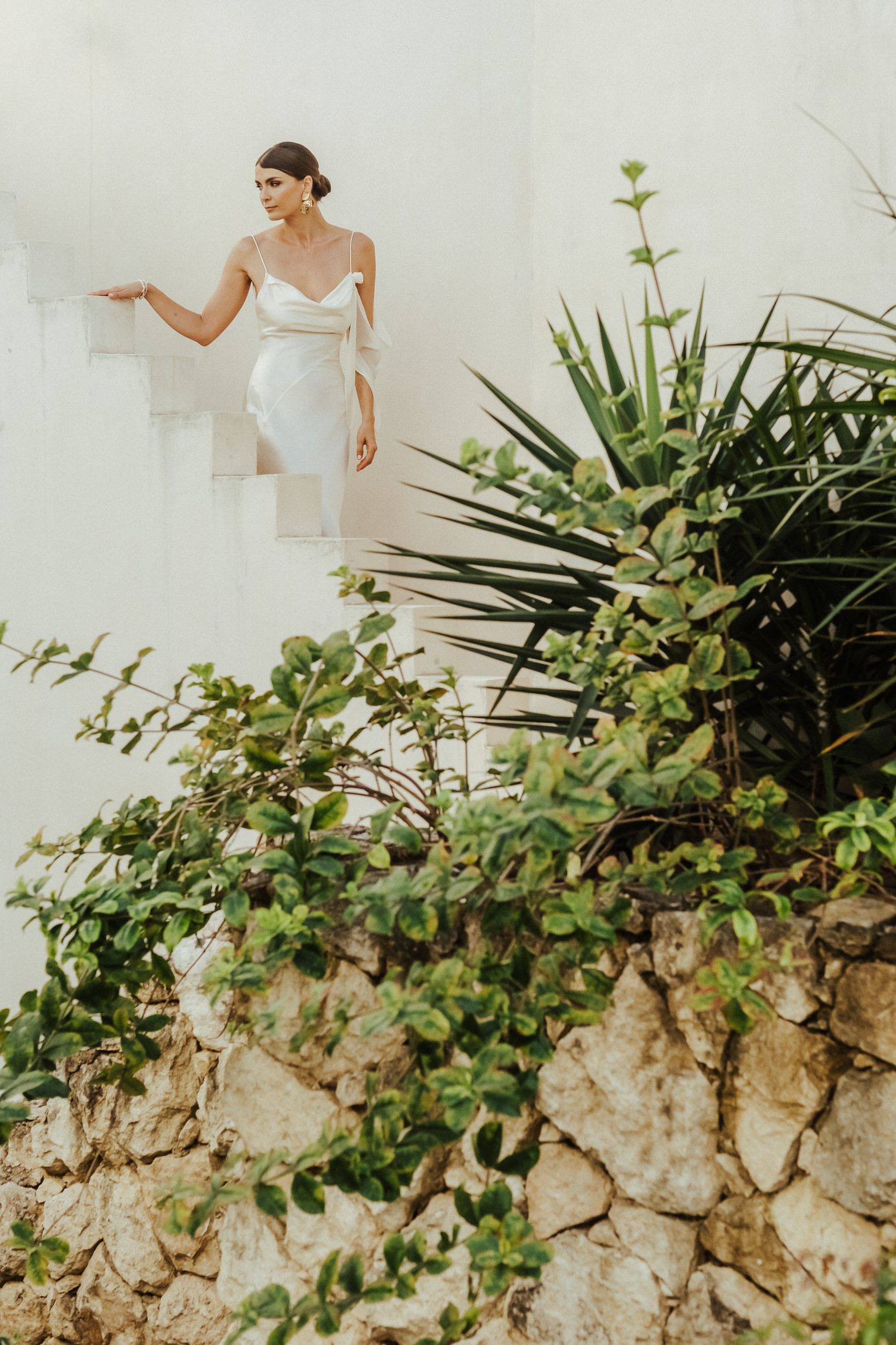 A figure in a white dress poses gracefully against a rustic stone wall with lush green foliage.