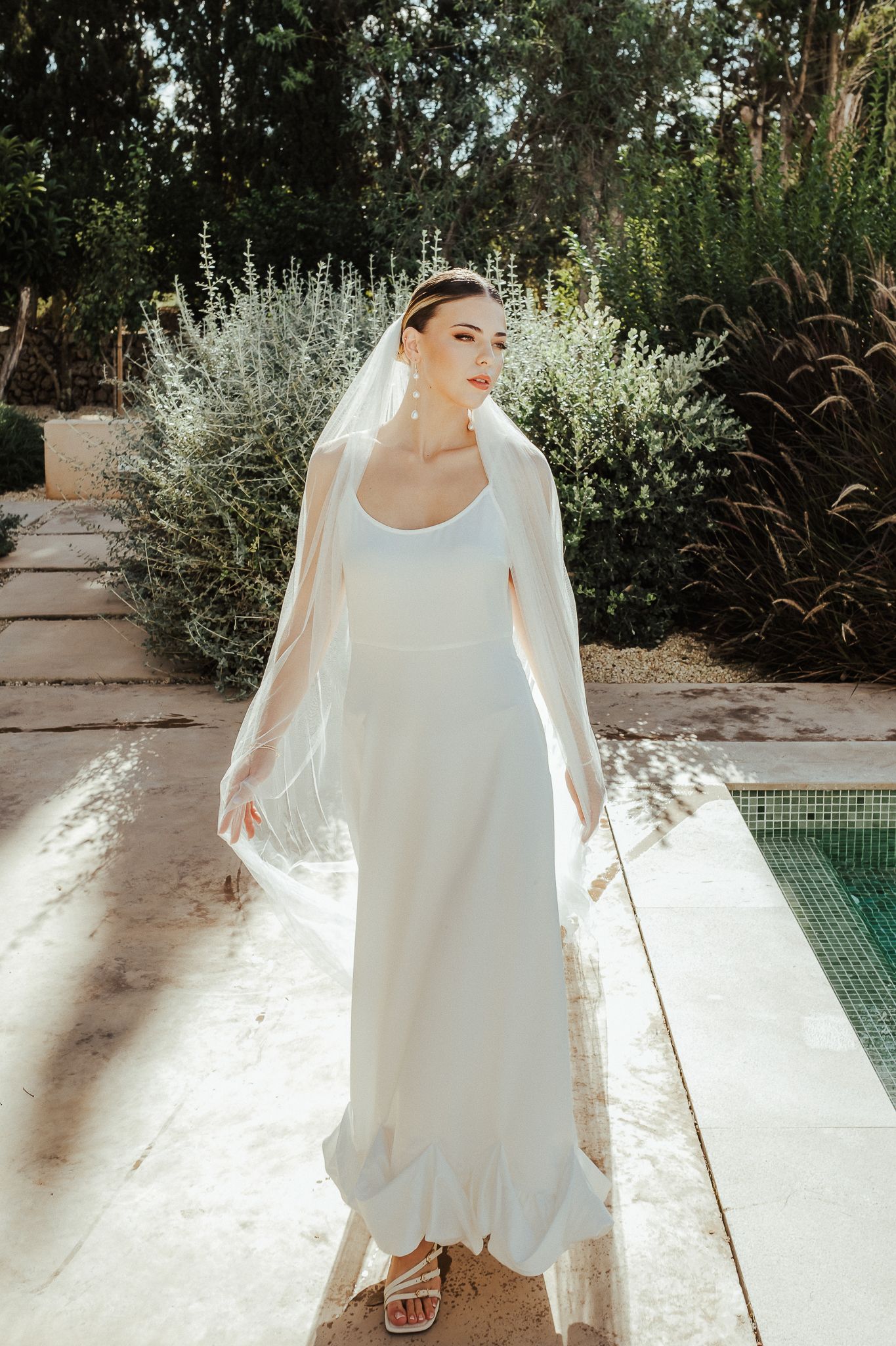 White wedding dress with flowy layers and tulle details photographed in sequence against a garden backdrop by a pool.