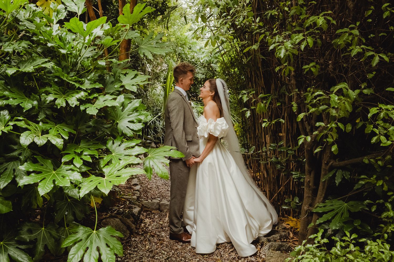 A couple in wedding attire share an intimate moment on a lush garden path surrounded by green foliage.