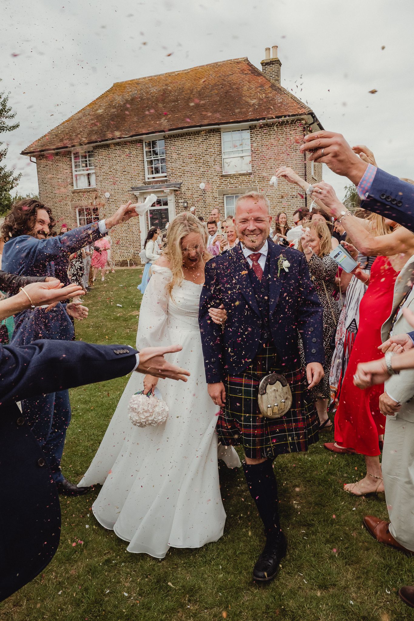 Wedding guests throw confetti over newlyweds outside a rustic brick building during a joyful celebration.