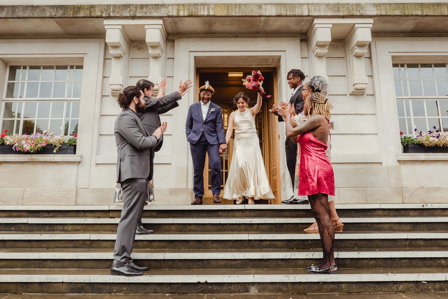 Wedding party poses for photos on grand stone steps of historic building during outdoor celebration.