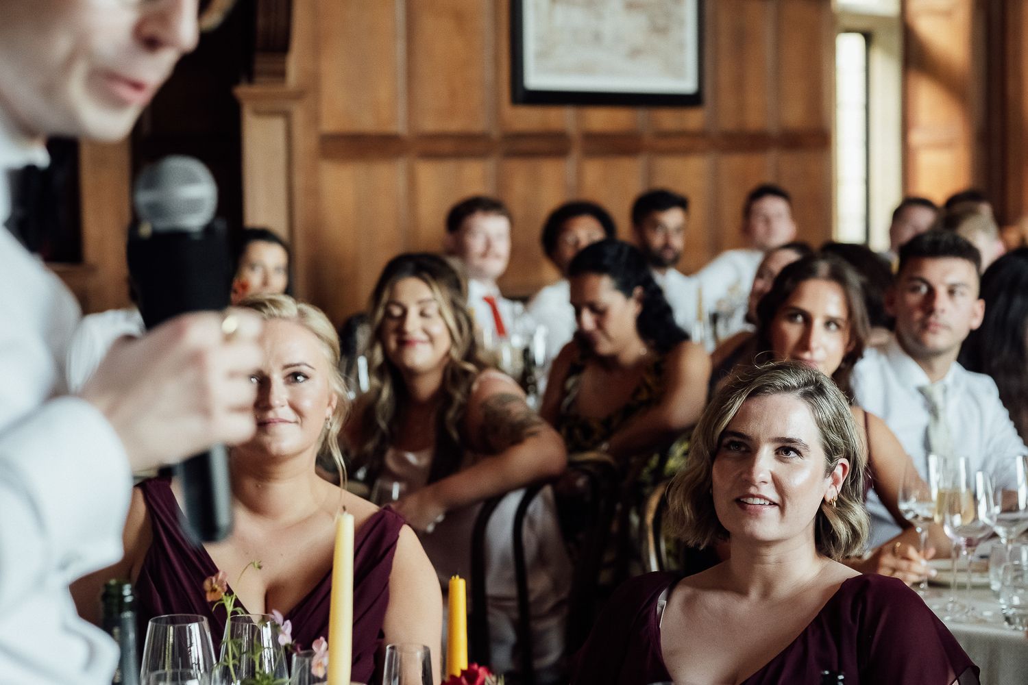 Bridesmaids in burgundy dresses smile listening to wedding speech at reception.