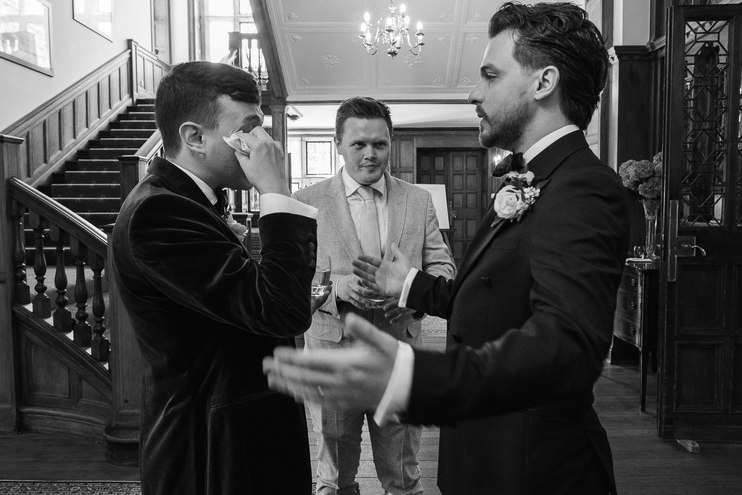 Black and white photo of three men in formal suits conversing at a wedding venue with elegant staircase.