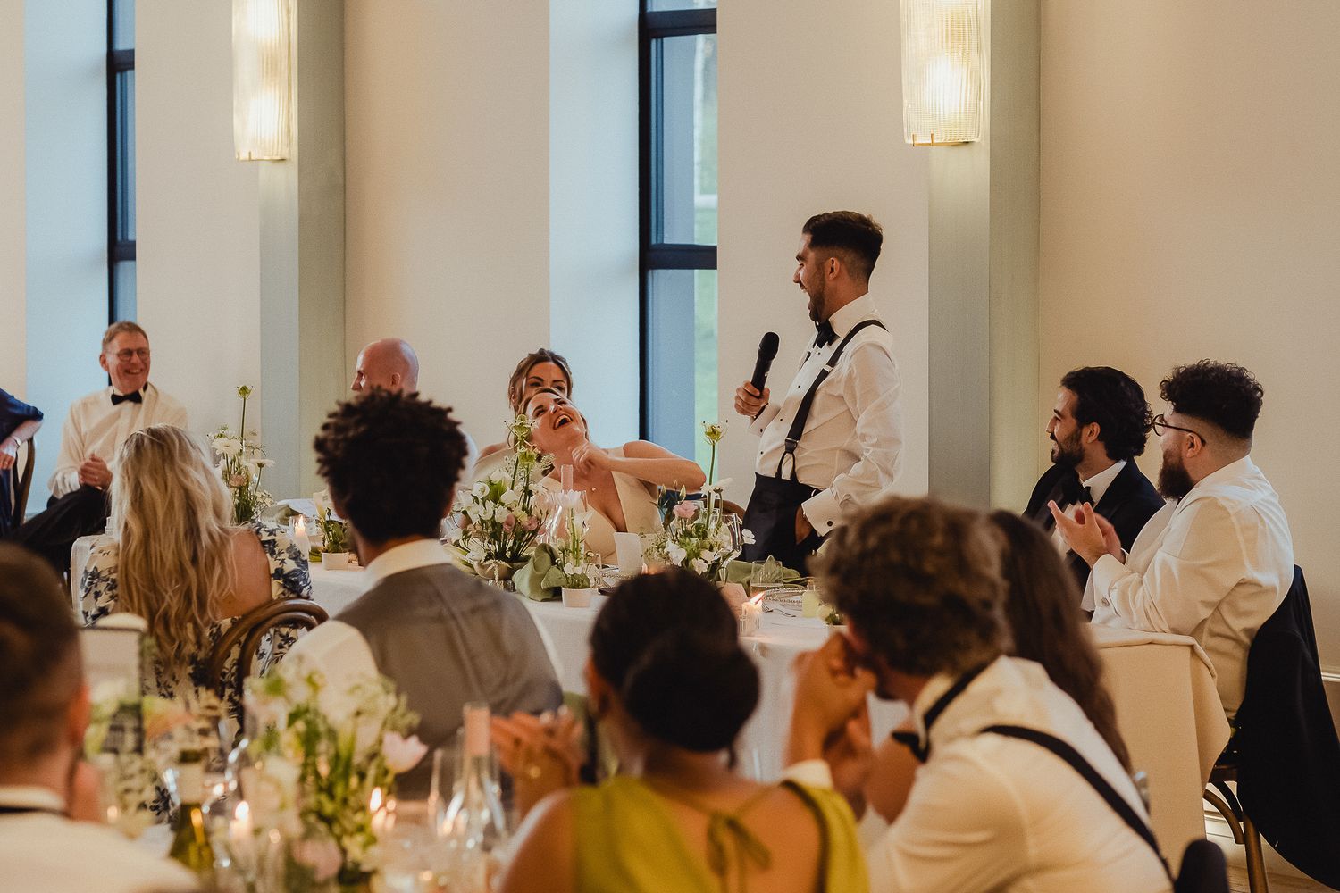 Wedding reception guests seated at decorated tables listen to someone giving a speech during dinner celebration.