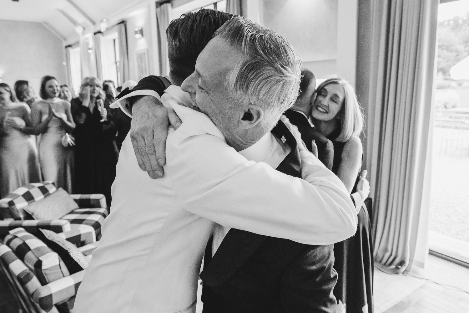 Black and white photo of an emotional embrace at a wedding celebration with guests watching in the background.