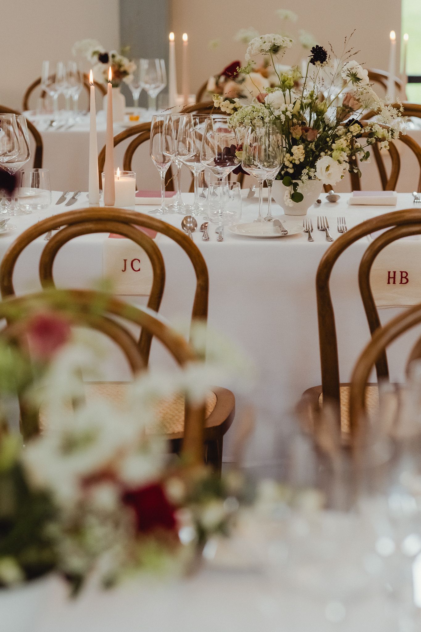 Elegant wedding reception tables with white linens, wooden chairs, and delicate floral centerpieces in a light-filled venue.