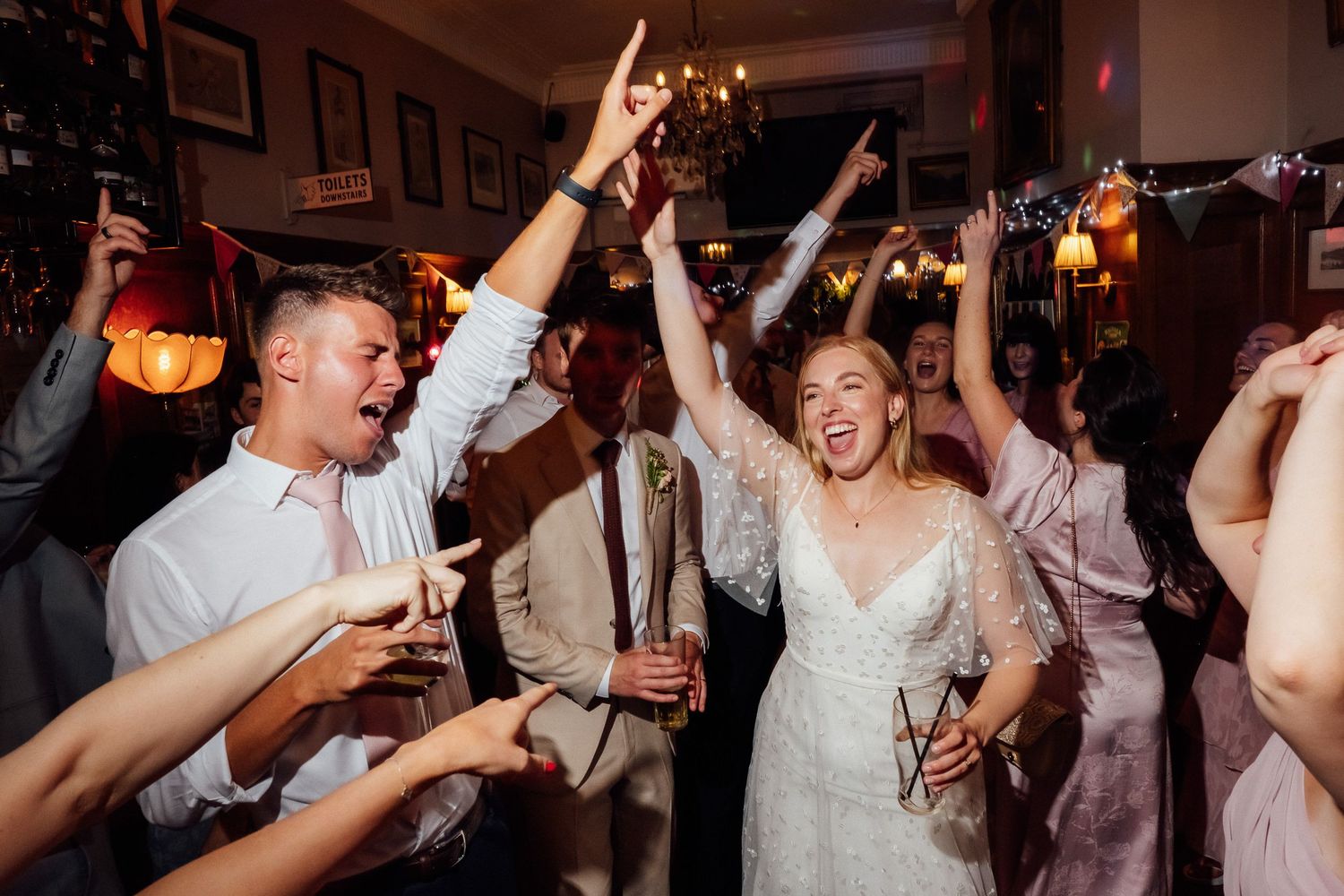 Bride in a white lace dress and groom in a white shirt dance joyfully with arms raised among cheering wedding guests at a lively reception.
