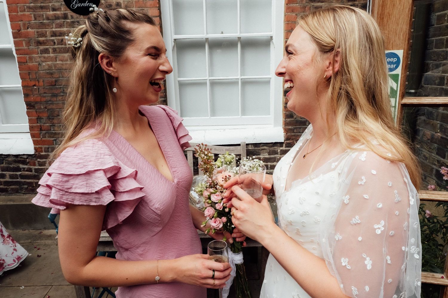 Two smiling women in pink and white dresses laugh together outside a brick building while holding a floral bouquet at a wedding celebration.