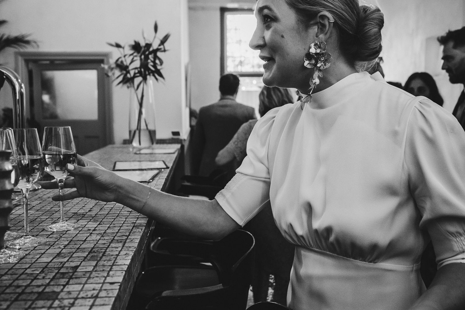 Black and white photo of an elegant party scene showing a guest at a bar with wine glasses and floral arrangements.
