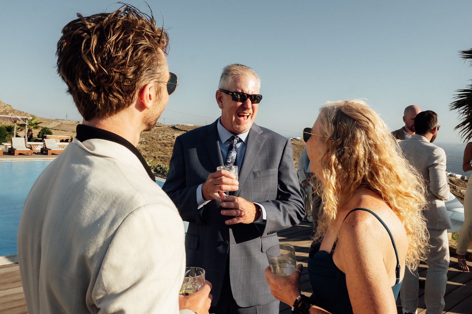 Three guests socializing at an outdoor wedding reception with scenic views, holding drinks and smiling.