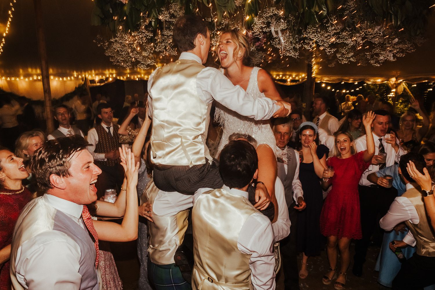 Wedding guests lifting a couple during an energetic dance party under string lights.