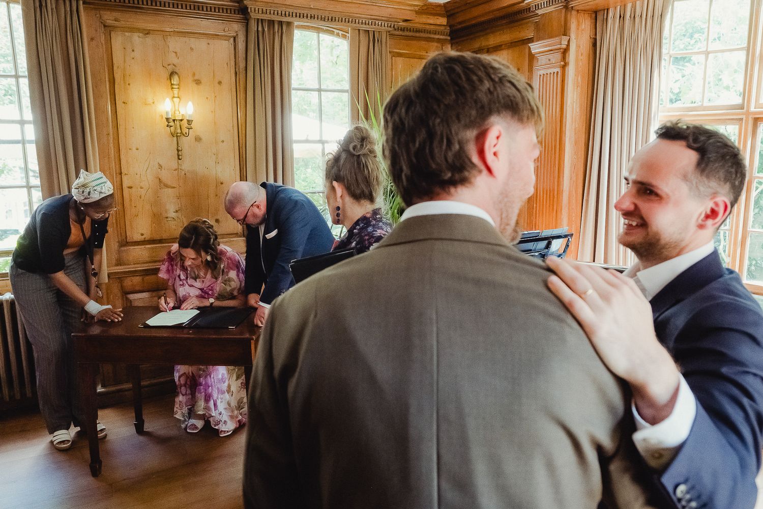 Two people in suits share a warm conversation at an indoor gathering while others mingle in the background.