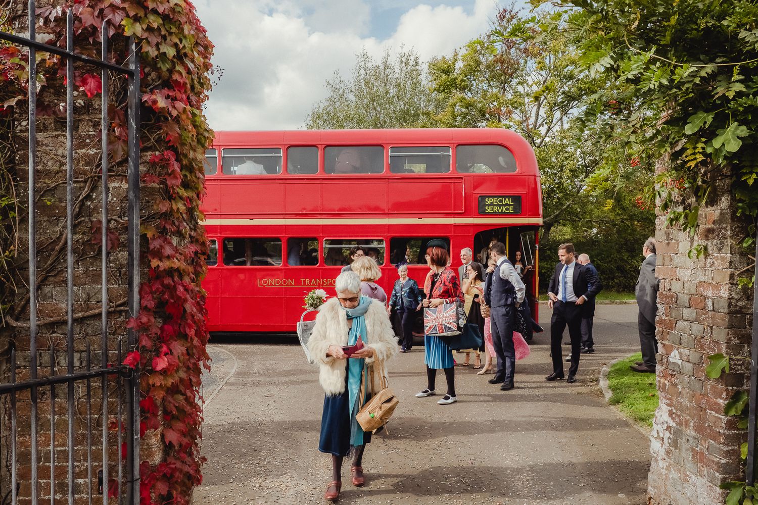Vintage red double decker bus parked on tree-lined path with visitors walking nearby in English countryside setting.