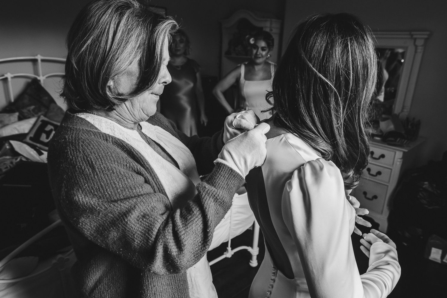 A black and white candid photo shows people getting ready in a dressing room with one person helping another with clothing.