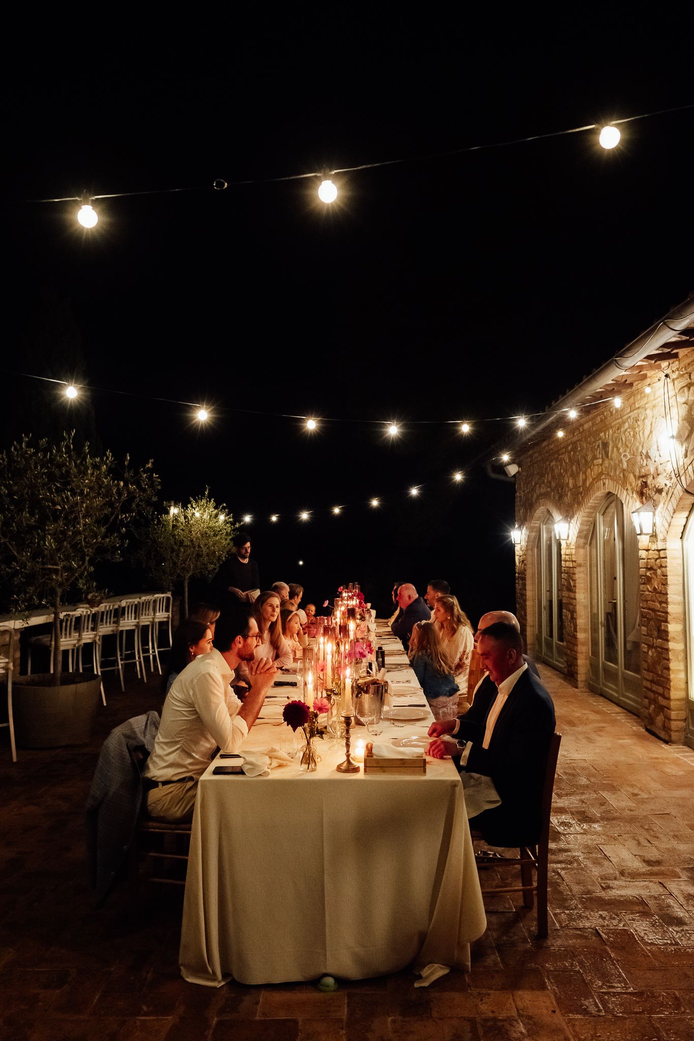Guests dine at a long candlelit table on a brick terrace at night under glowing string lights beside a rustic stone building.