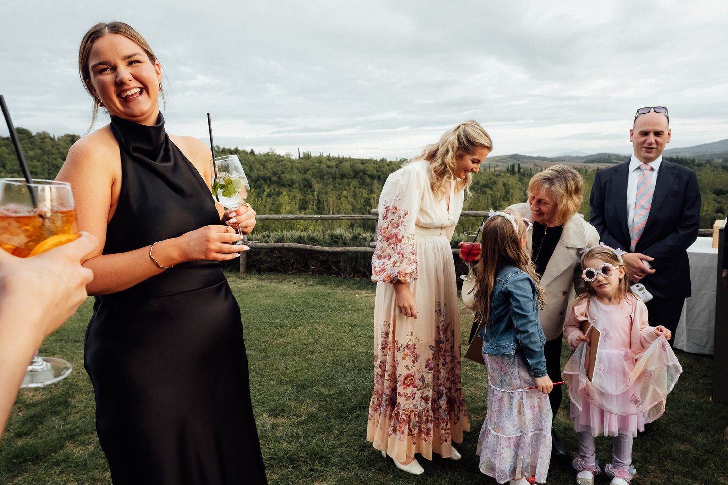 Laughing woman in a black dress holds a cocktail at an outdoor wedding reception while guests and two young girls mingle on a grassy lawn.