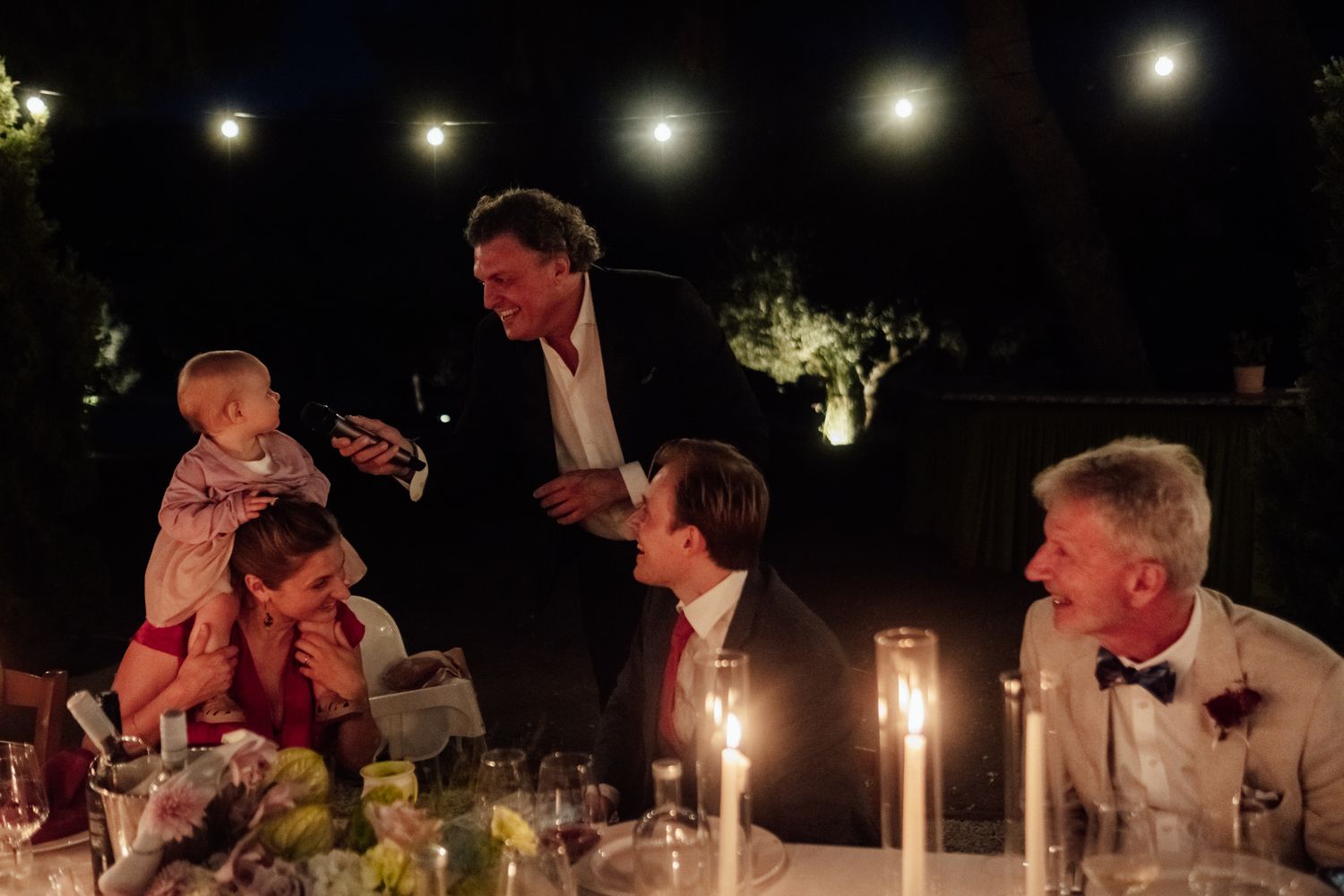 Guests in formal attire laugh and play with a baby around a candlelit table at an outdoor evening dinner under glowing string lights.