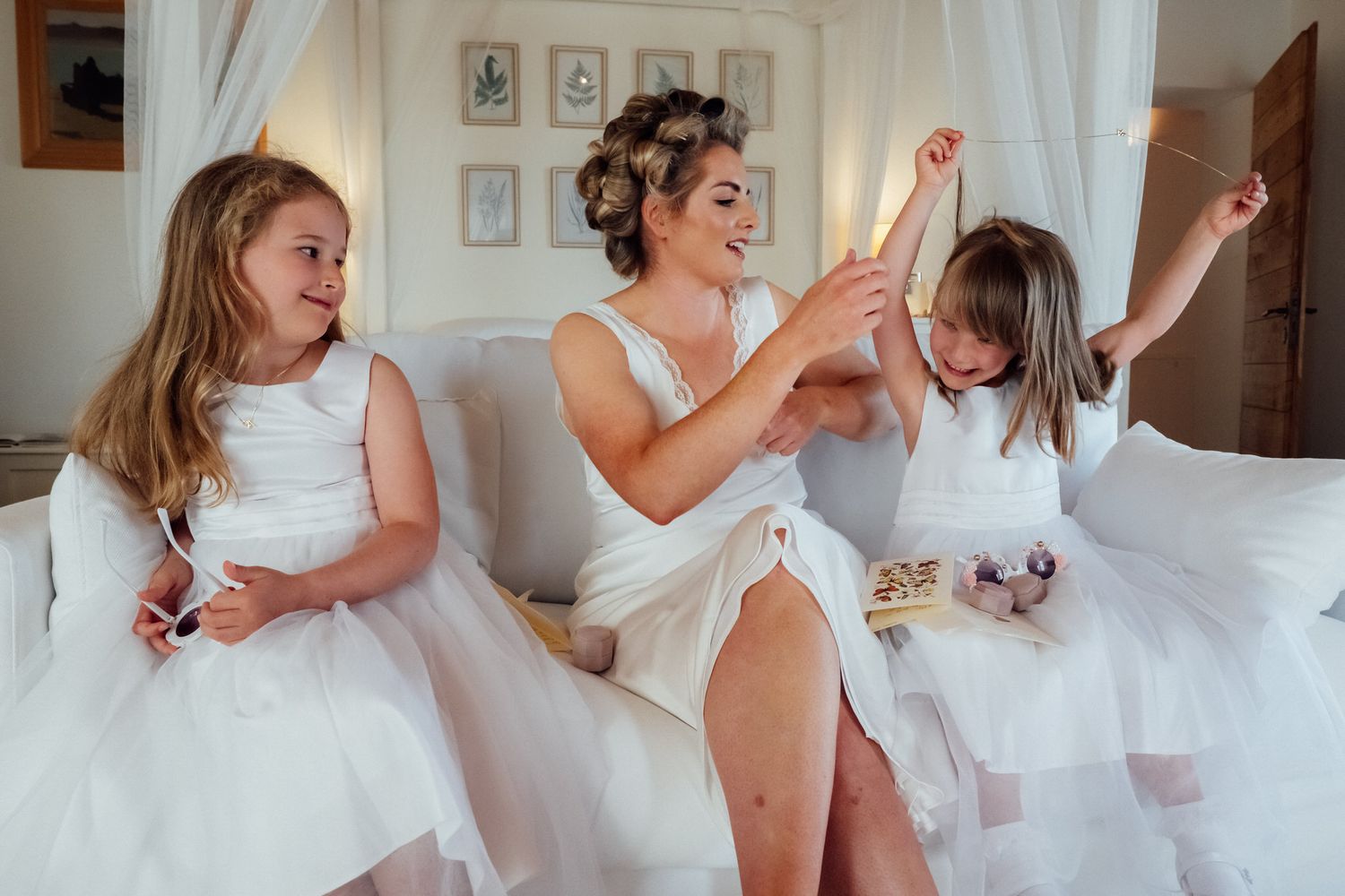 A bride in a white dress laughs with two flower girls in white tulle dresses while sitting on a bed in a bright, curtained room.
