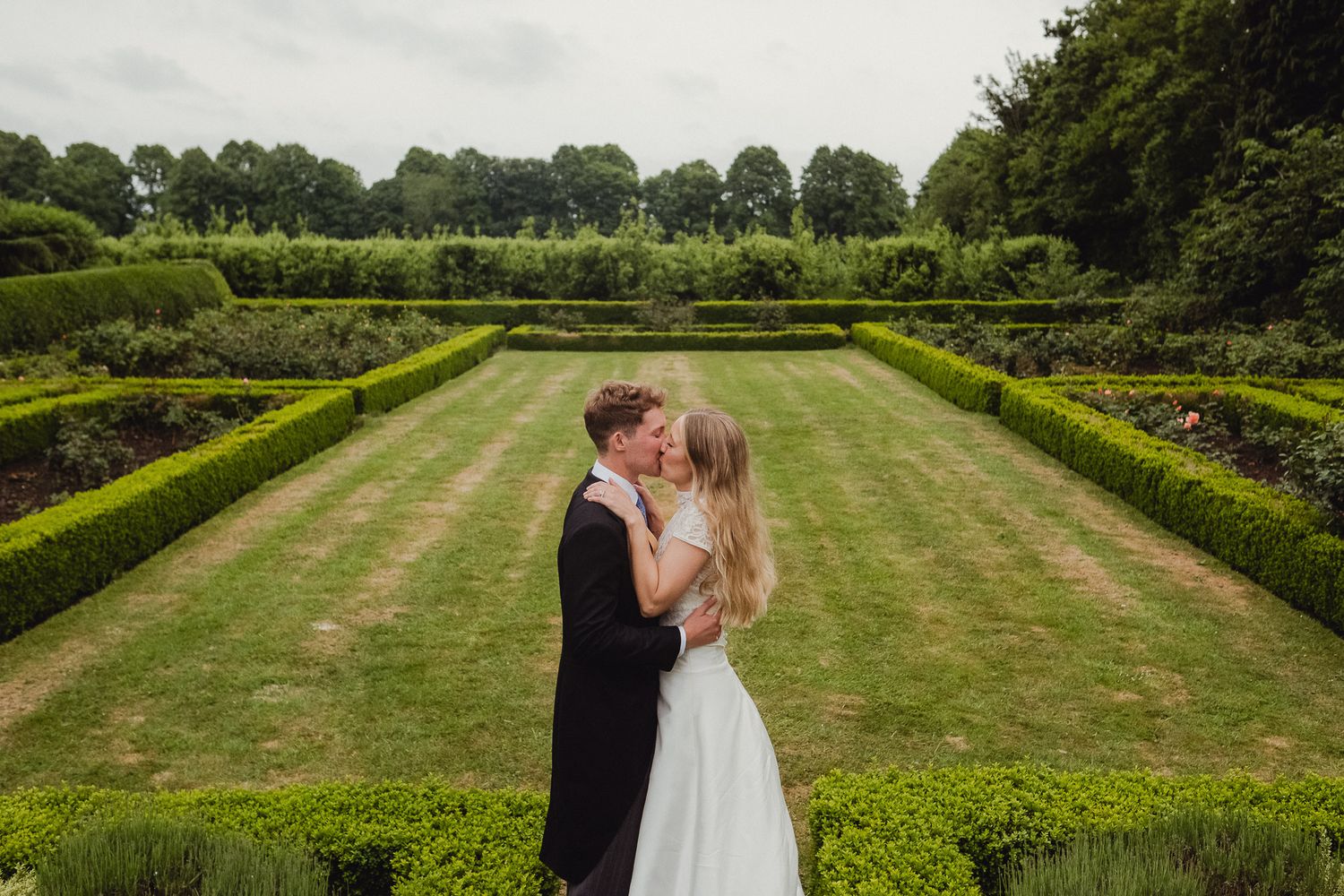 A couple shares an intimate moment in a romantic garden setting surrounded by manicured hedges and green lawn.
