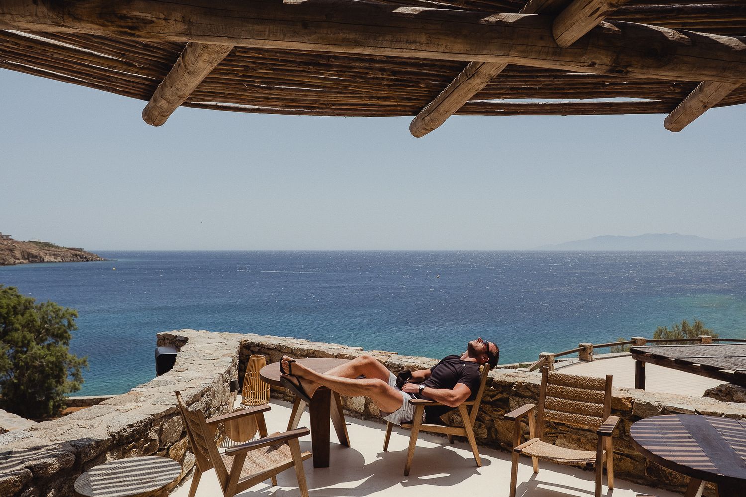 Rustic wooden outdoor beach lounge chairs and umbrellas overlooking a serene Mediterranean coastline on a sunny day.