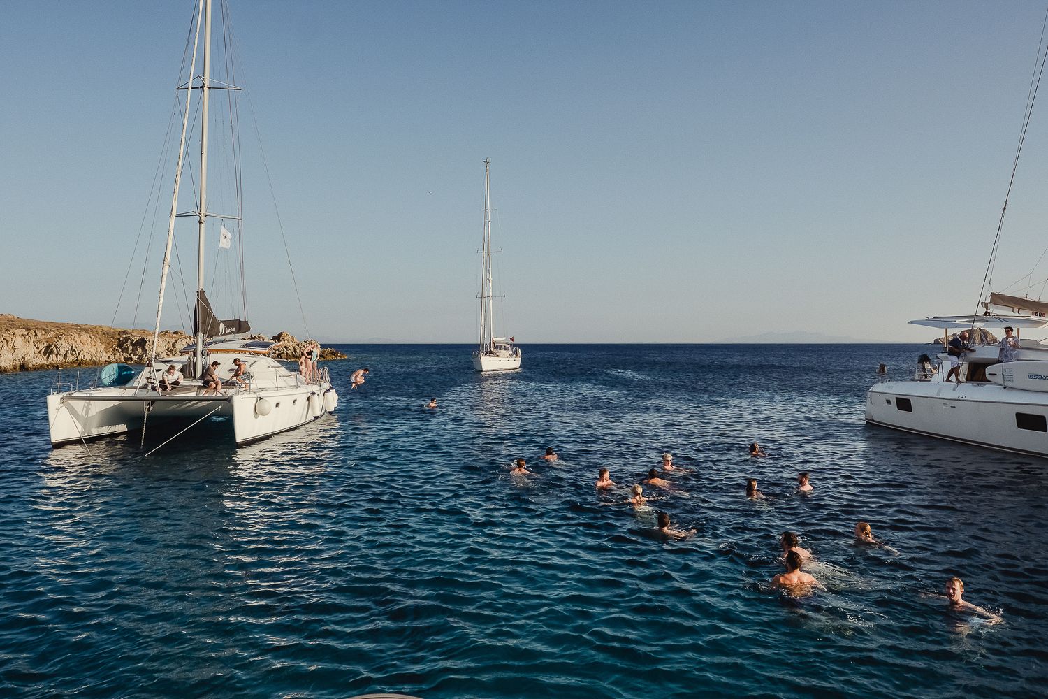 Sailboats and catamarans anchored in crystal clear blue Mediterranean waters with rocky coastline visible in background.
