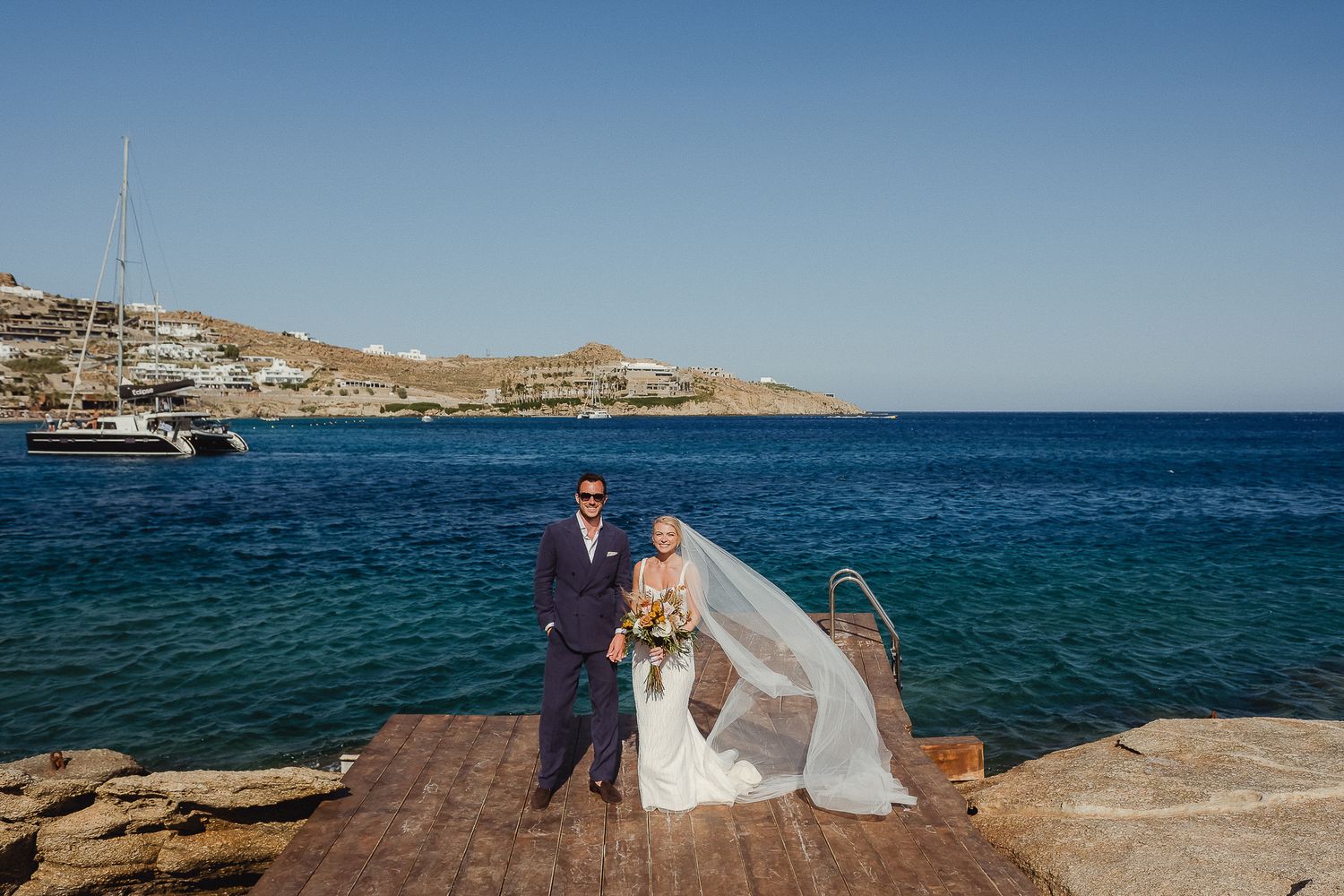 A seaside wedding portrait captured on a rustic wooden pier with blue waters and boats in the background.