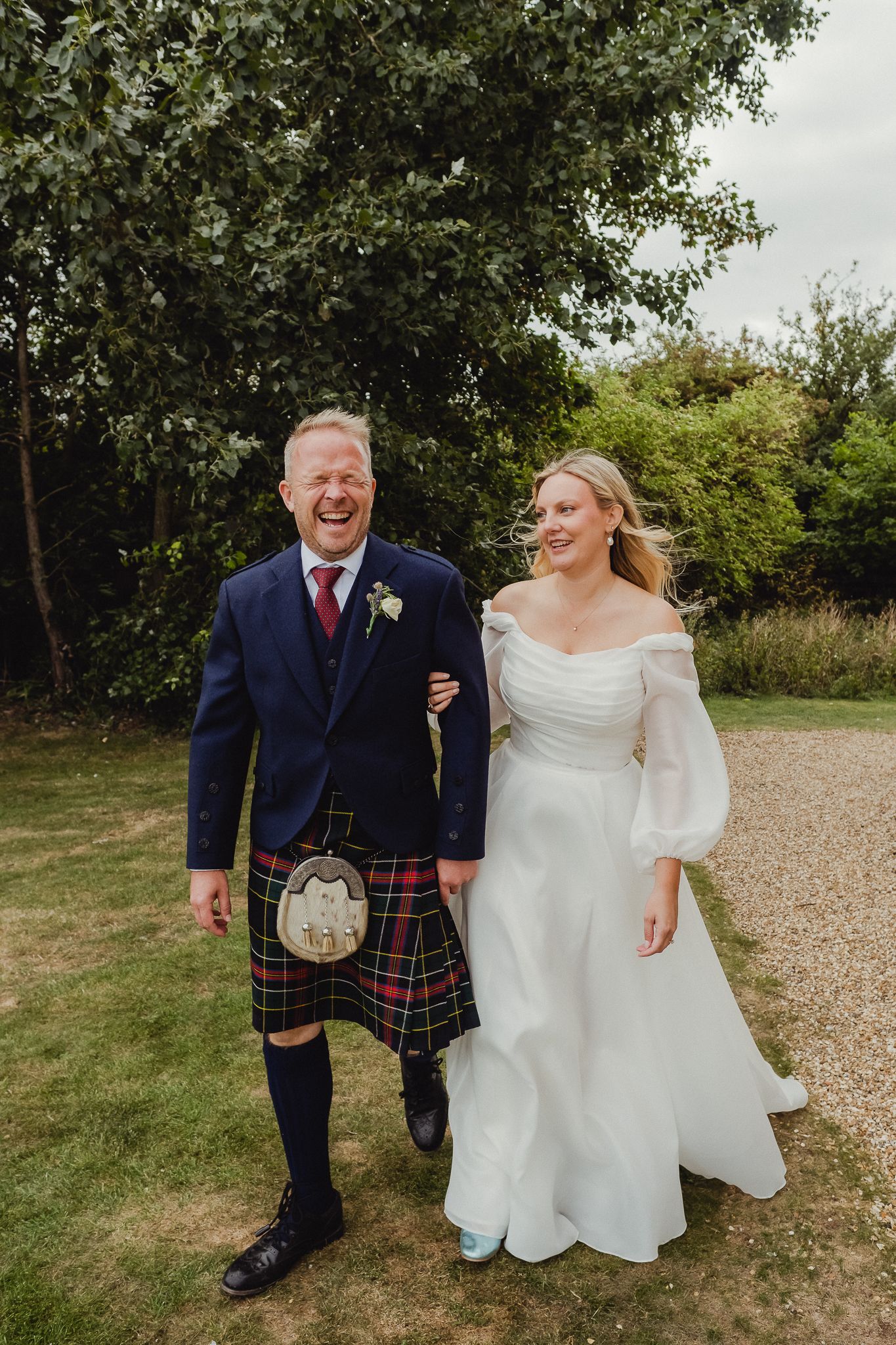 A couple walks together outdoors on their wedding day, with the groom wearing a traditional Scottish kilt and navy jacket.