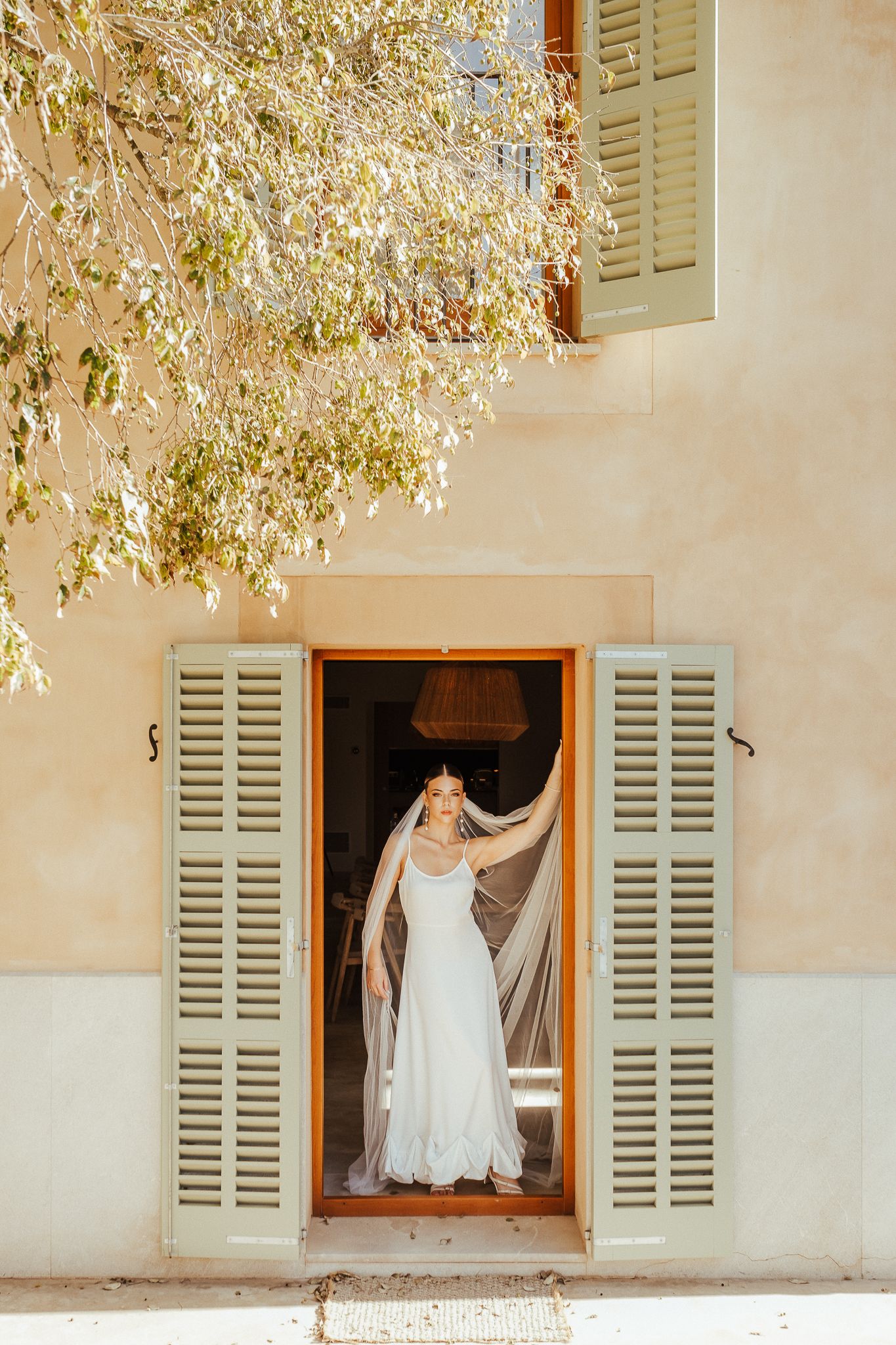 A white dress billows in the breeze through a rustic window with pale shutters against a Mediterranean style wall.