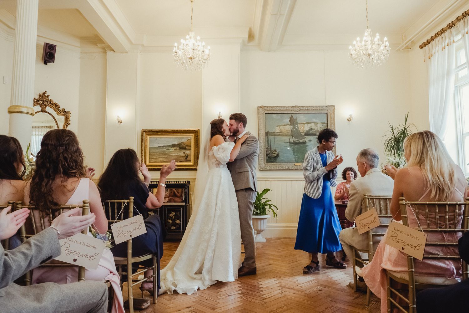 Wedding party members gather in an elegant room with chandeliers and hardwood floors during a formal celebration.