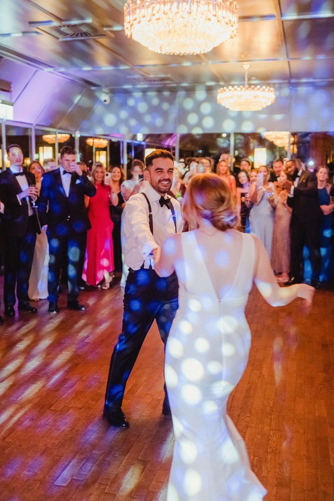 Dancing couples celebrate on a wooden dance floor under crystal chandeliers at an elegant wedding reception.