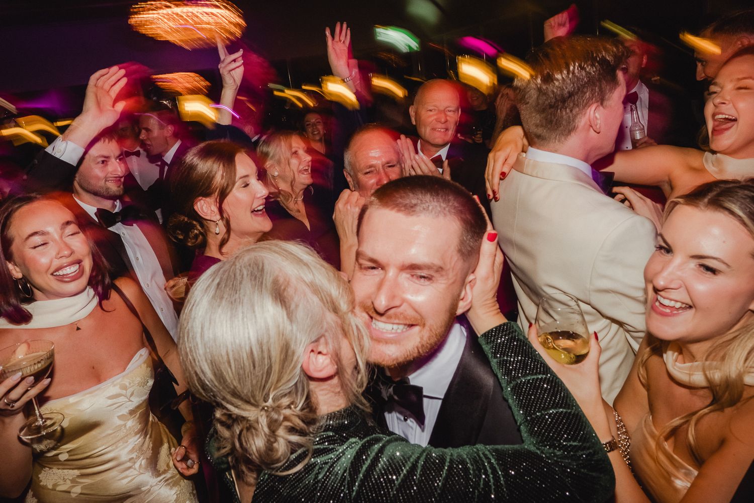 Energetic crowd of party guests dancing and celebrating at a festive reception with colorful lighting.
