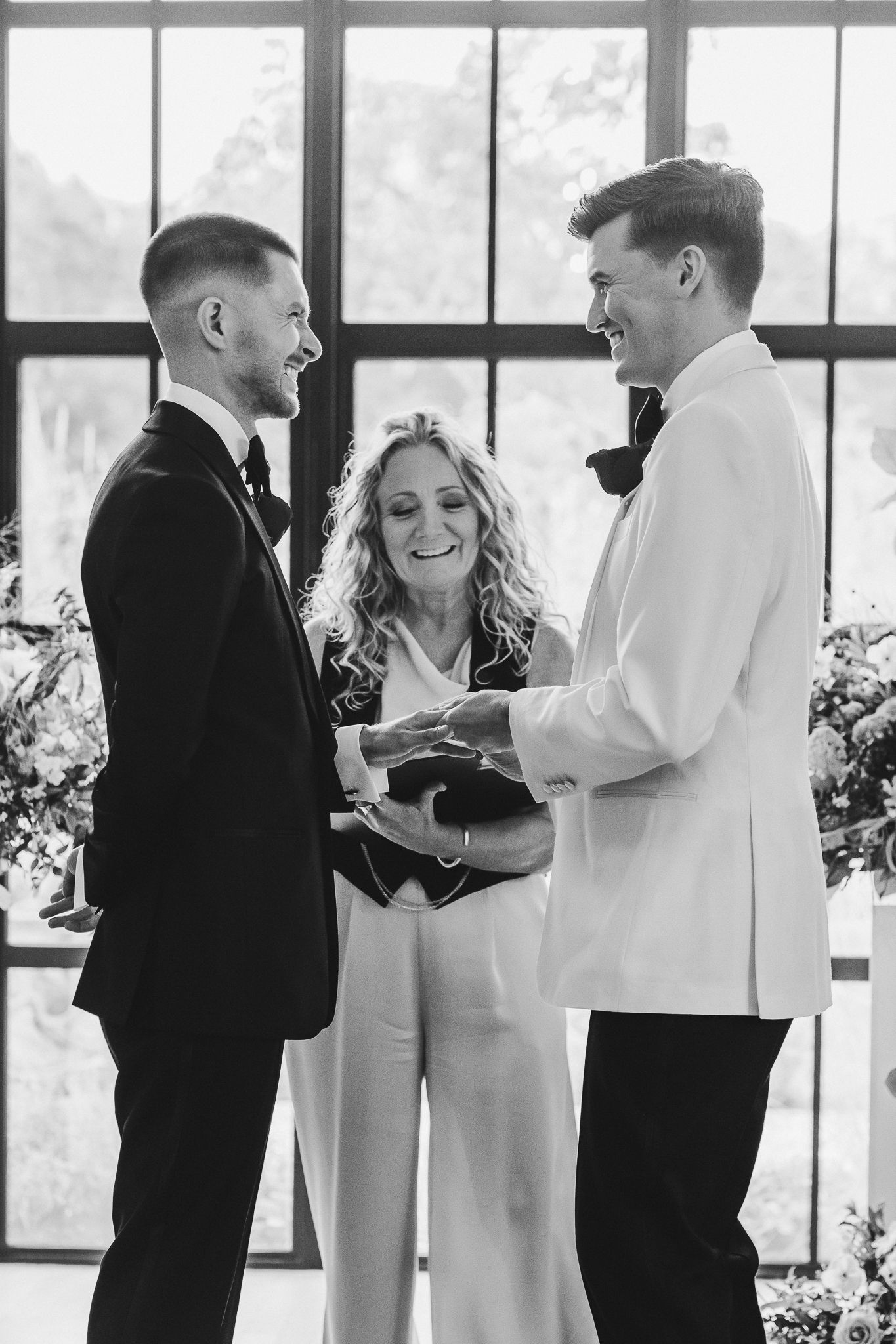 A black and white photo shows wedding guests mingling and laughing at a reception by large windows.
