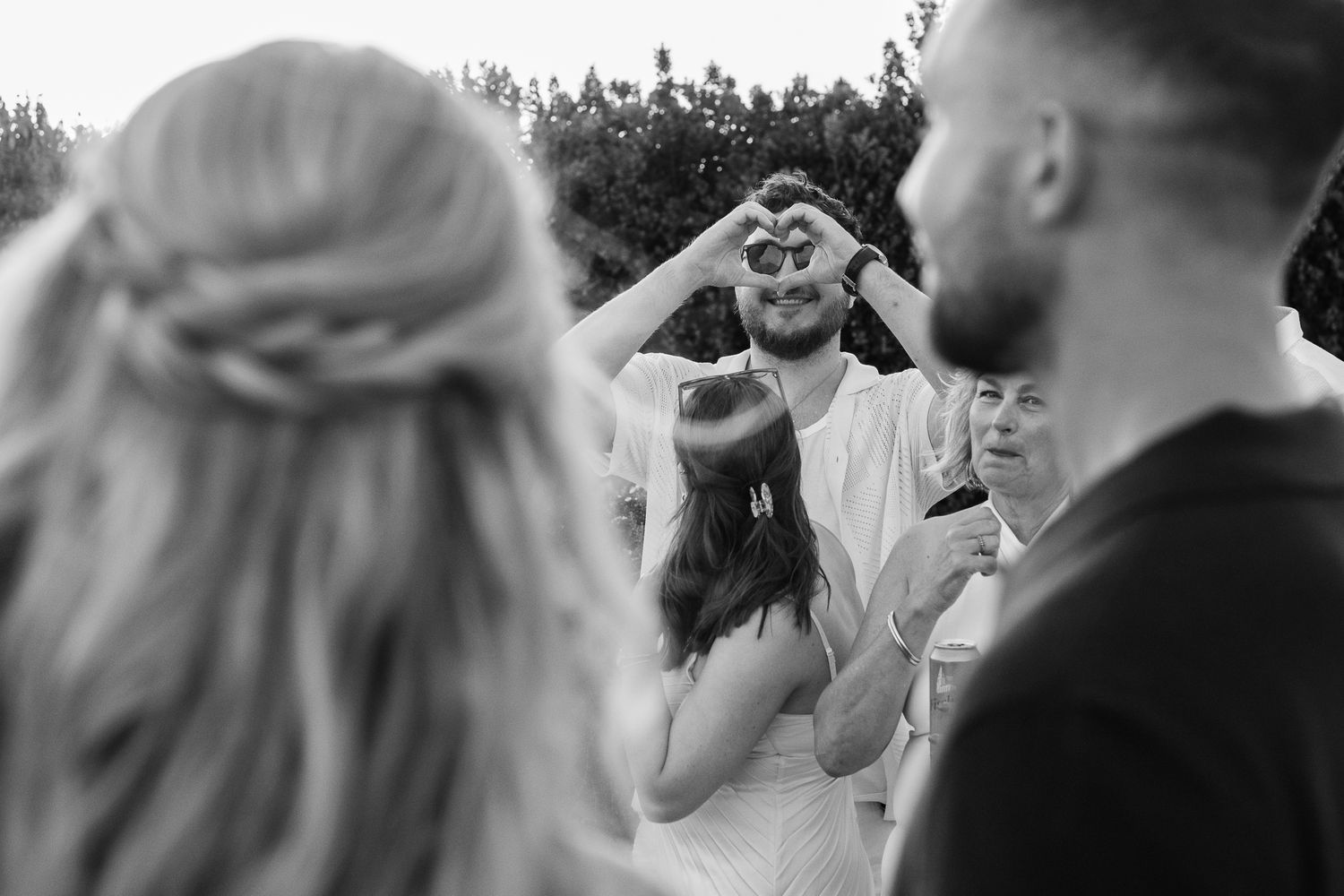 Black and white wedding photo of groom making heart shape with hands framing bride at outdoor ceremony.