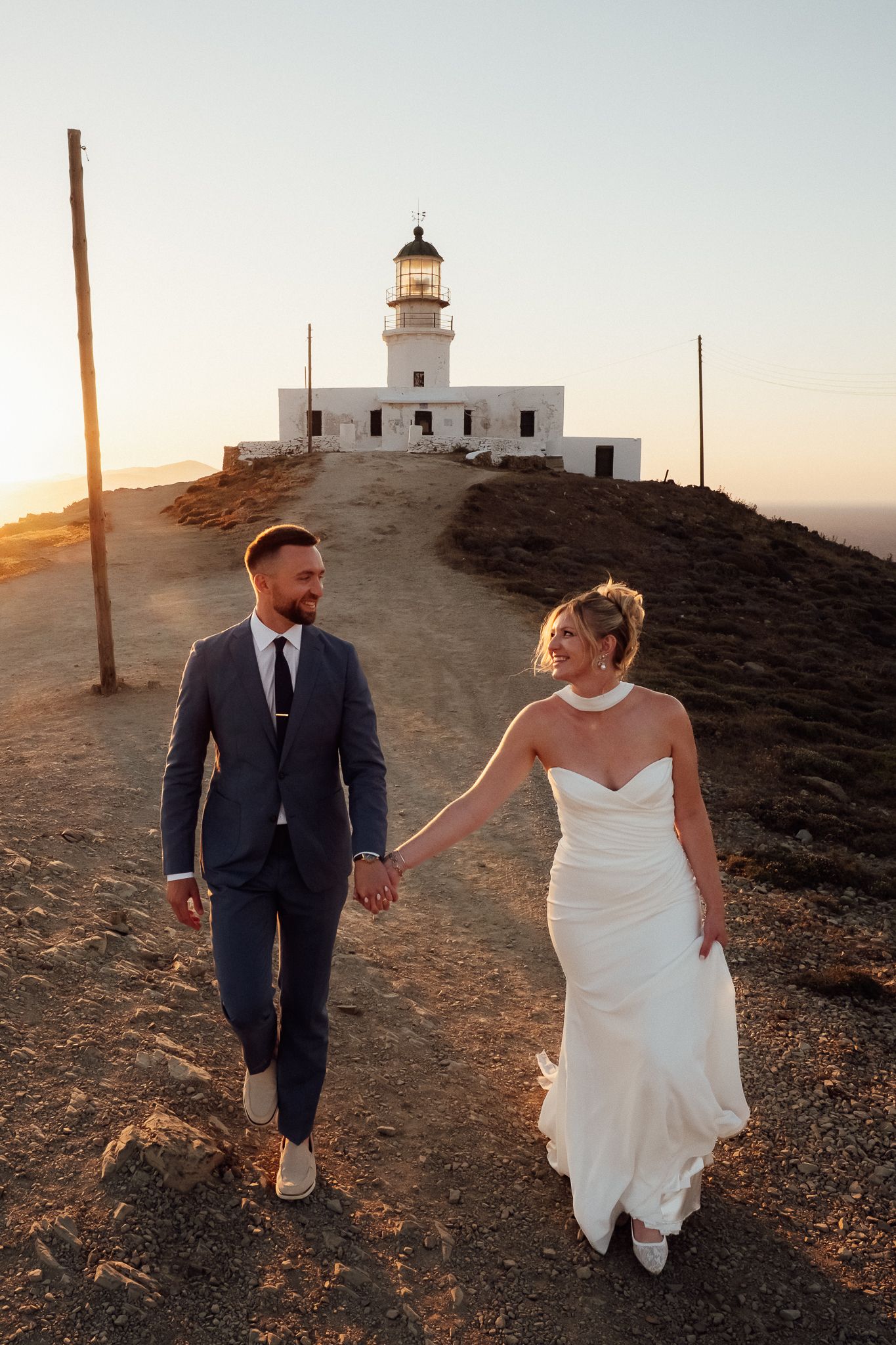 Bride and groom hold hands walking from a lighthouse at sunset during their Mykonos elopement.