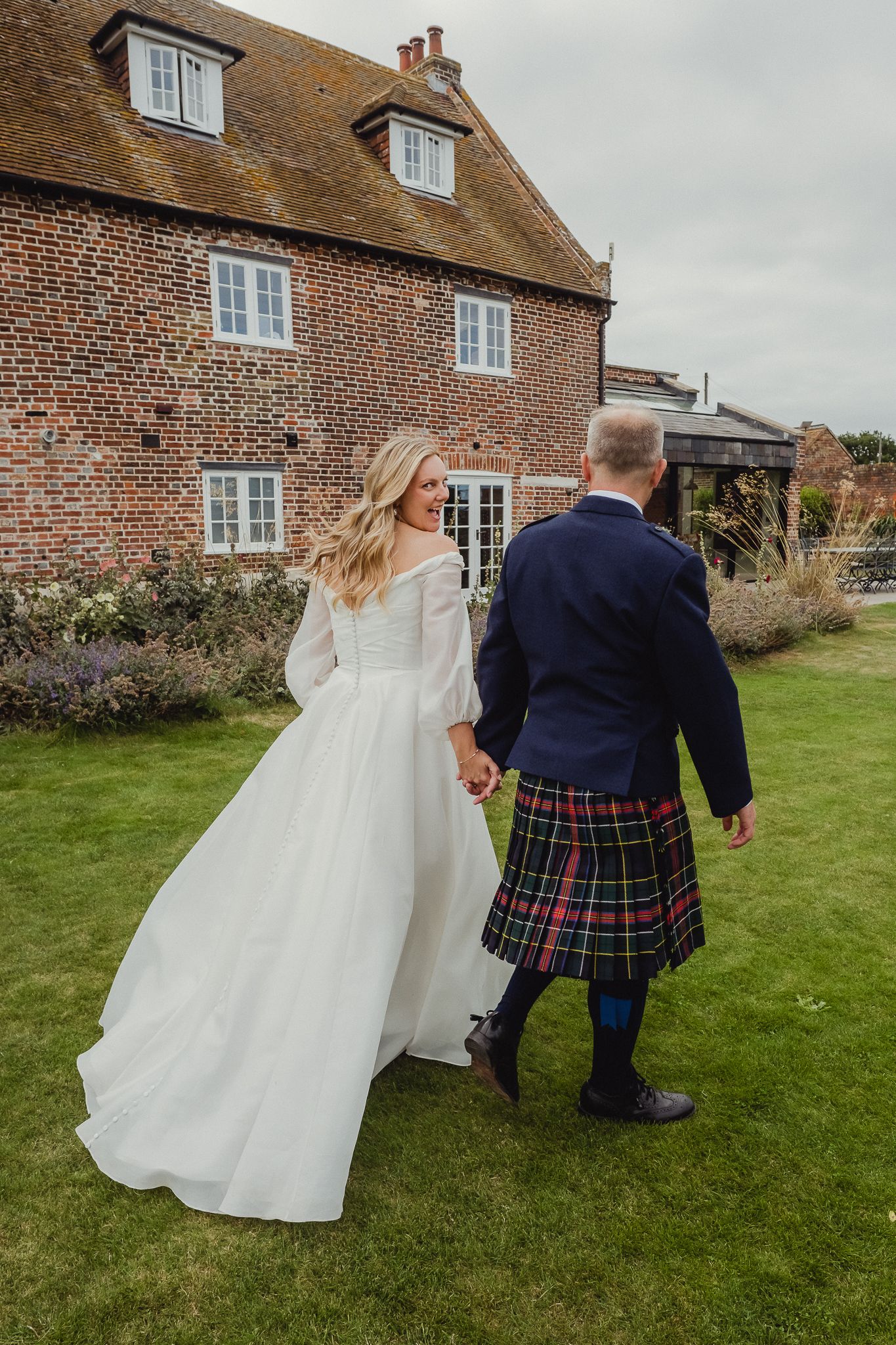 A couple in wedding attire walking away from the camera across a green lawn beside a brick country house.