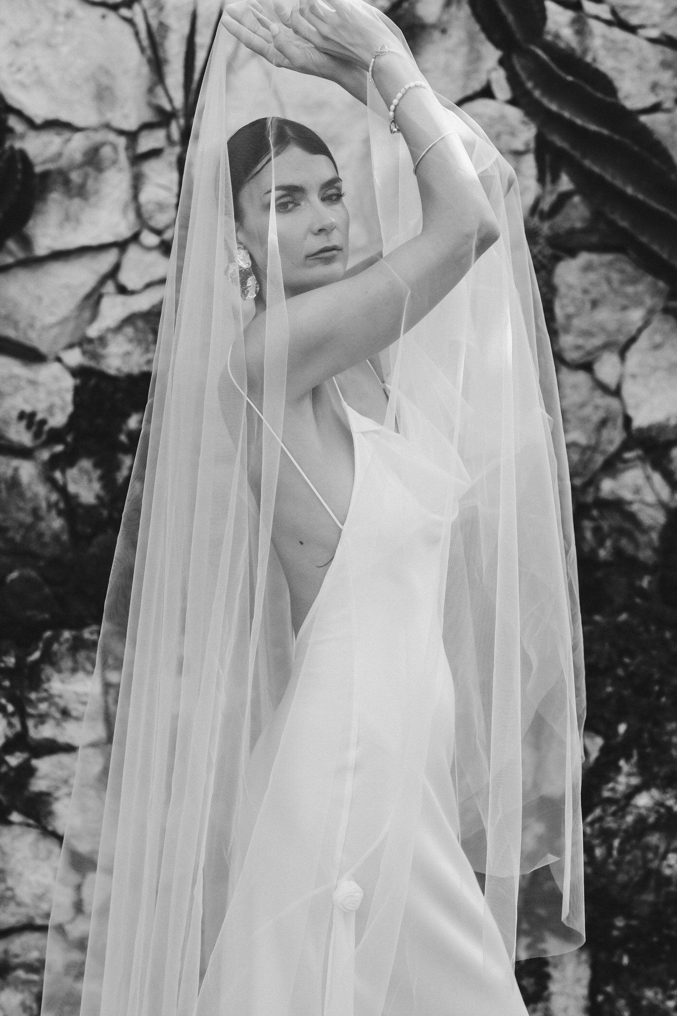 A bride in a white wedding dress and flowing veil poses against a textured stone wall in an artistic series.