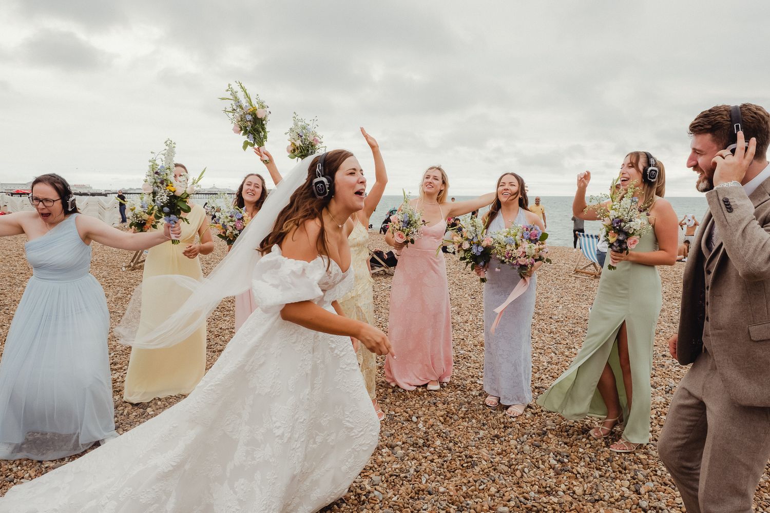 A joyful wedding party celebrates on a beach with bridesmaids in pastel dresses and the bride in white.